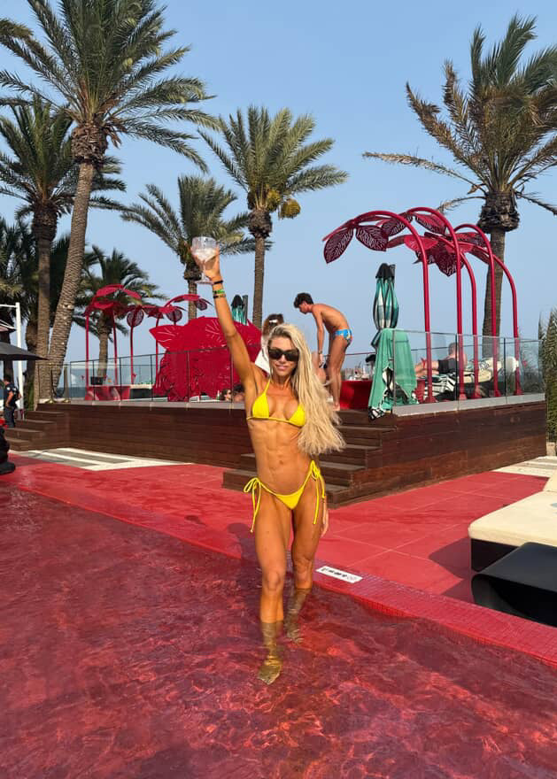 Woman in a yellow bikini standing on a red pool deck with palm trees and a poolside area in the background.